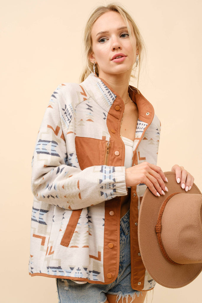 A woman wearing a beige jacket with an Aztec-inspired geometric pattern and earthy brown accents, featuring a chest pocket and snap buttons.