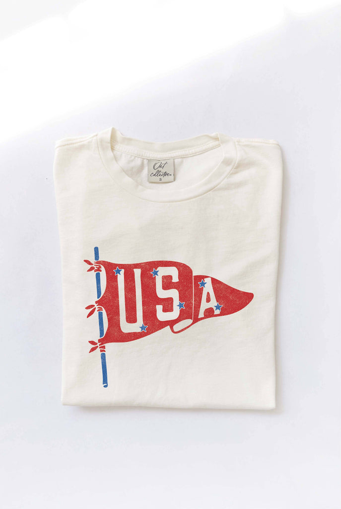 Woman wearing a cream-colored T-shirt with a vintage red USA pennant graphic, styled with beige high-waisted pants.