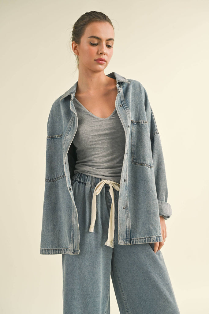 Model in an oversized denim jacket with front pockets. First image: jacket buttoned, paired with white shorts. Second image: jacket open, over a grey tank and denim pants.