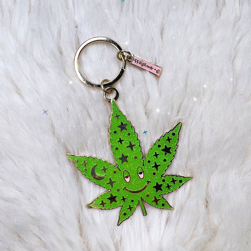 A vibrant green enamel weed leaf keychain with a happy face and a starry pattern, including a moon and stars, against a white fluffy background.