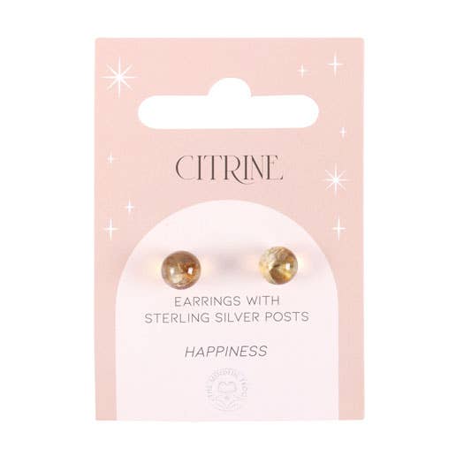 A pair of citrine crystal stud earrings displayed on a matching backing card, held in a hand with a serene background.