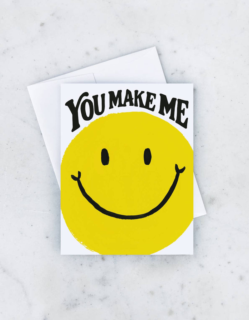 A greeting card with a large yellow smiley face and the words “You Make Me” in bold, playful lettering.