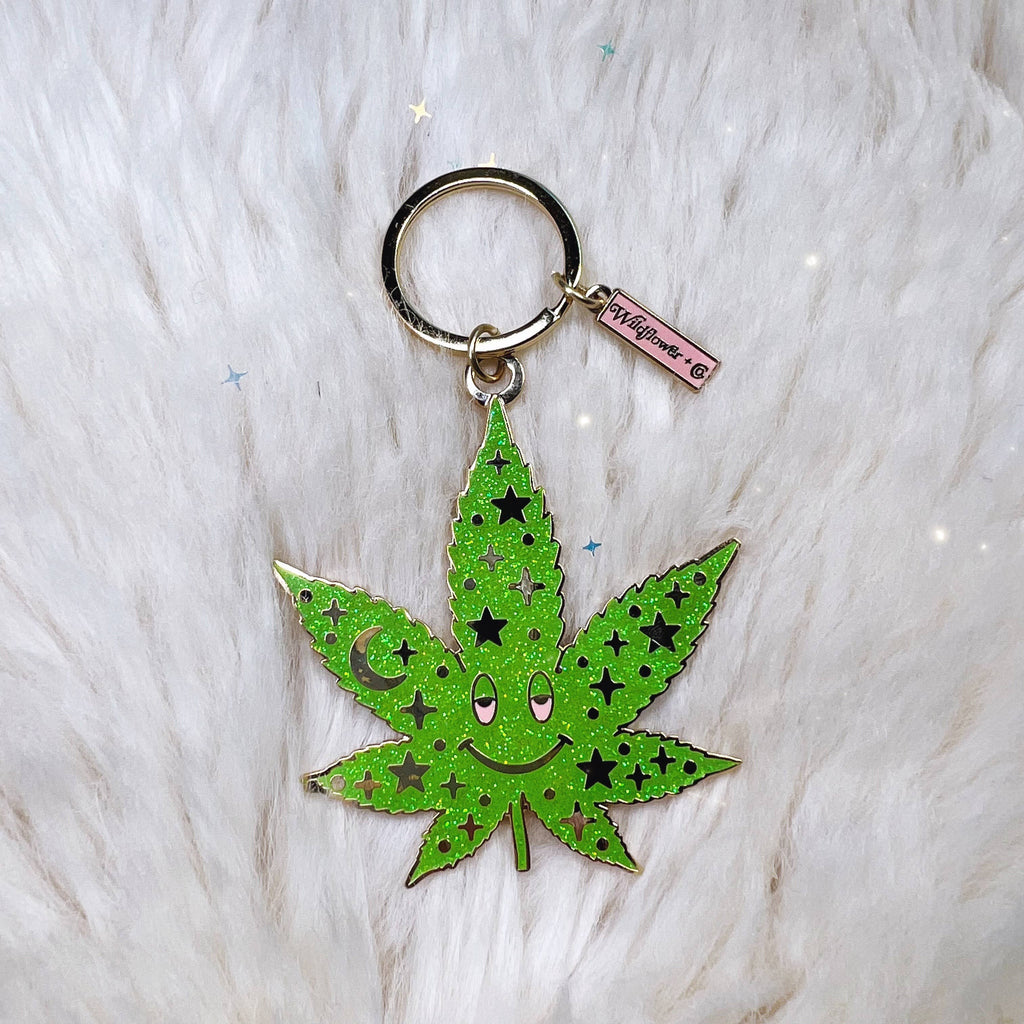 A vibrant green enamel weed leaf keychain with a happy face and a starry pattern, including a moon and stars, against a white fluffy background.