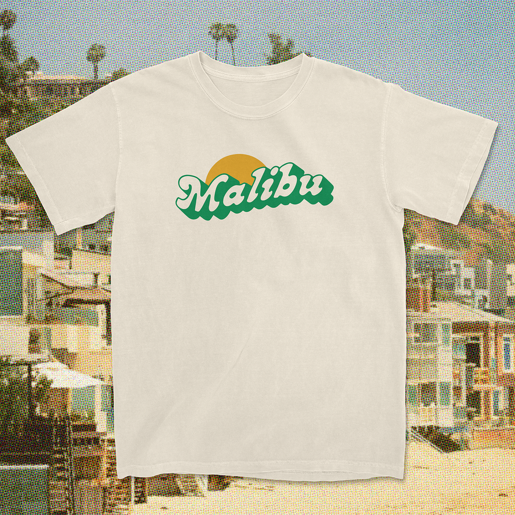 White "Malibu" T-shirt with green text and sun graphic, set against a scenic Malibu beach background.