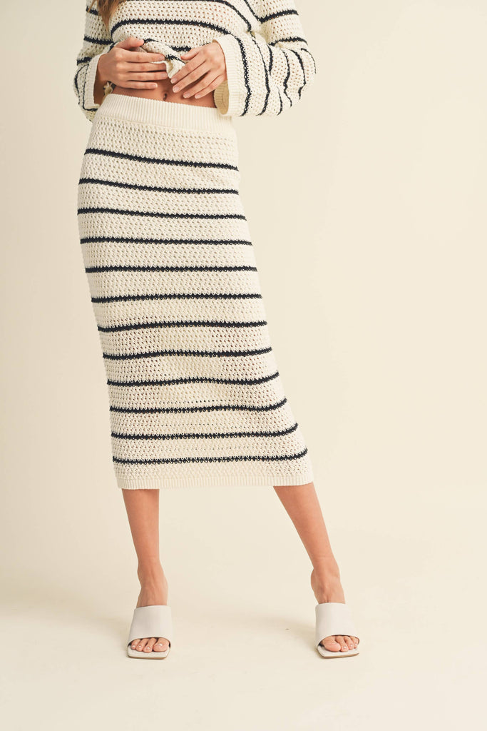 Woman wearing a high-waisted, ivory and black striped knit midi skirt with slide sandals.