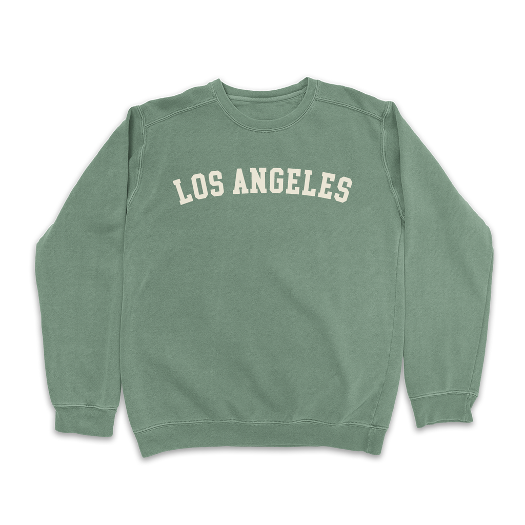 Light blue "Los Angeles" sweatshirt on a sunny palm-lined street background.
