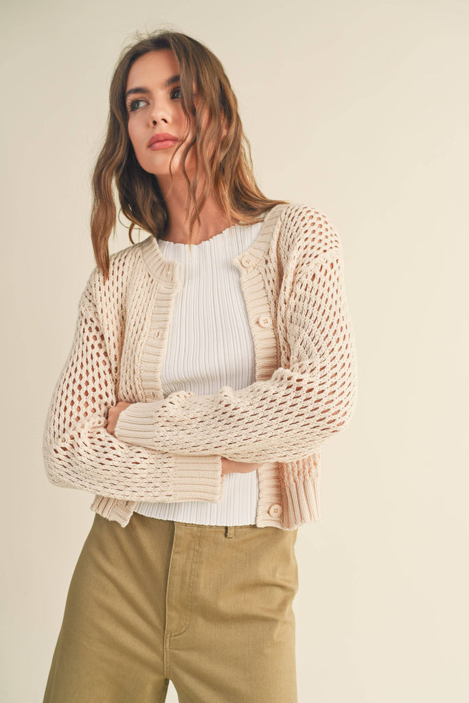 Model in a cream open-knit cardigan. cardigan over a white top and khaki pants. cardigan slightly open, paired with the same outfit.