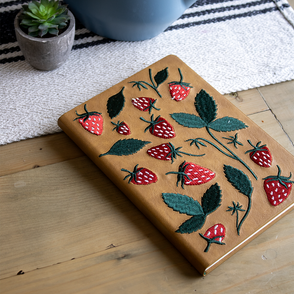 Embroidered journal with a strawberry design, featuring high-quality paper and durable binding, placed on a wooden table.