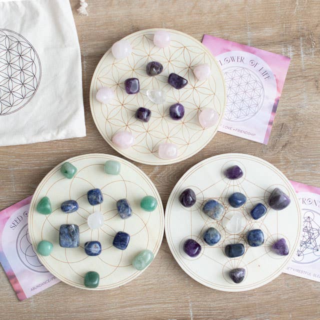 A Flower of Life crystal grid set featuring a wooden grid with a geometric flower of life pattern, arranged with various crystals including rose quartz, amethyst, and clear quartz. Surrounded by candles, flowers, and additional crystals on a black background.