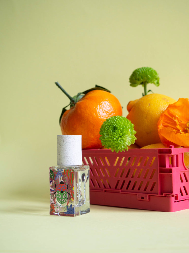 Maison Matine Nature Insolente perfume bottle with colorful leaf and beetle illustrations, placed on a vibrant yellow fabric and surrounded by green foliage.