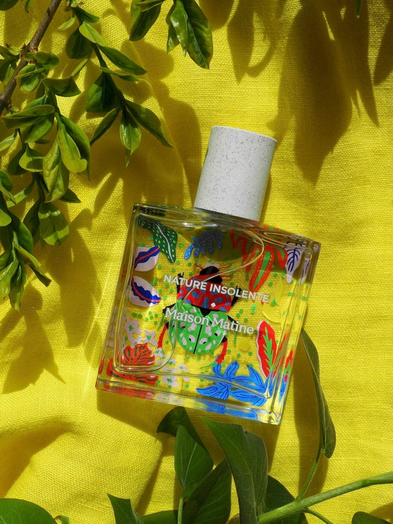 Maison Matine Nature Insolente perfume bottle with colorful leaf and beetle illustrations, placed on a vibrant yellow fabric and surrounded by green foliage.