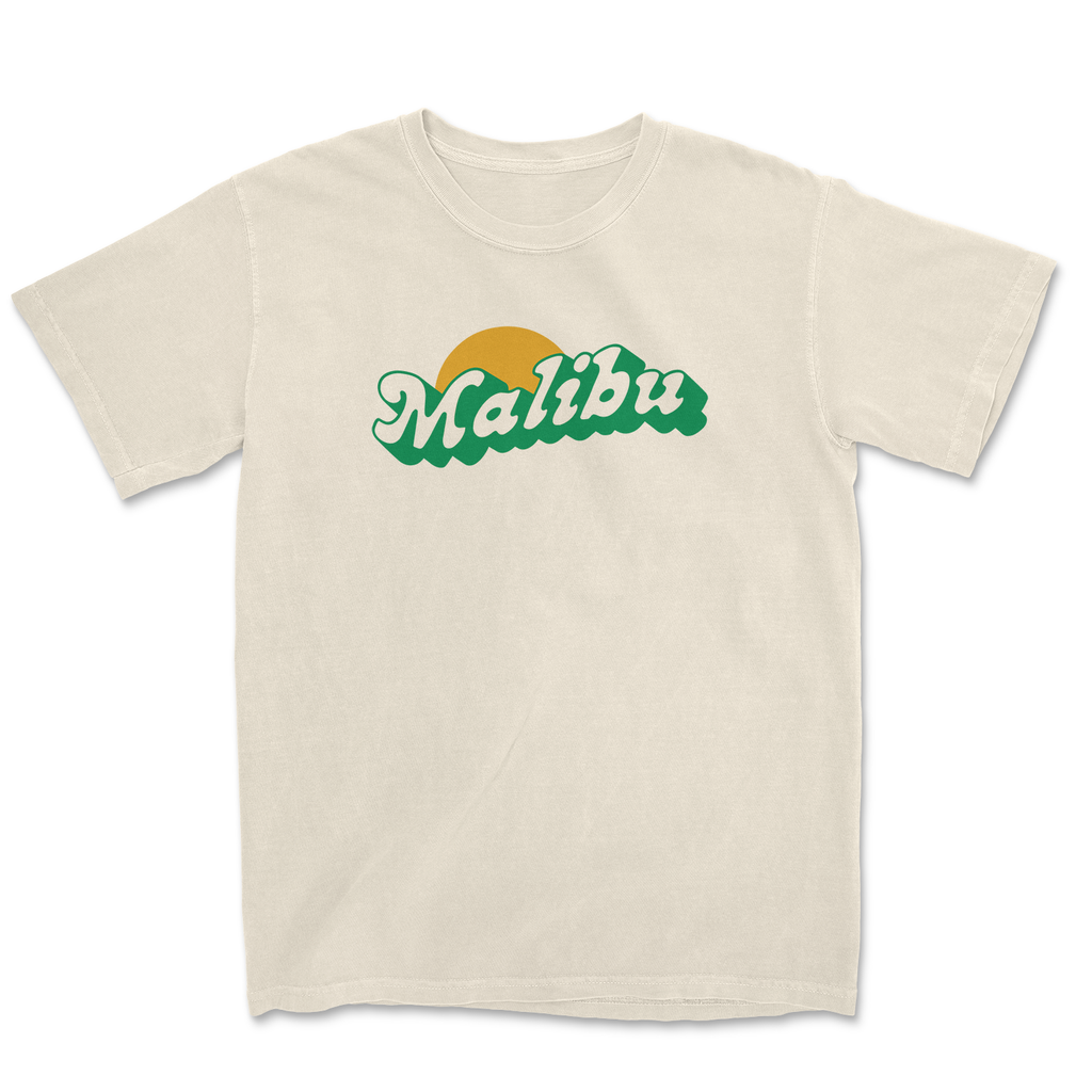 White "Malibu" T-shirt with green text and sun graphic, set against a scenic Malibu beach background.