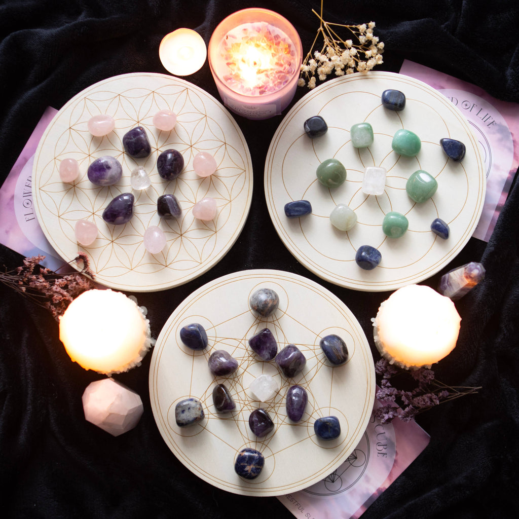 A Flower of Life crystal grid set featuring a wooden grid with a geometric flower of life pattern, arranged with various crystals including rose quartz, amethyst, and clear quartz. Surrounded by candles, flowers, and additional crystals on a black background.