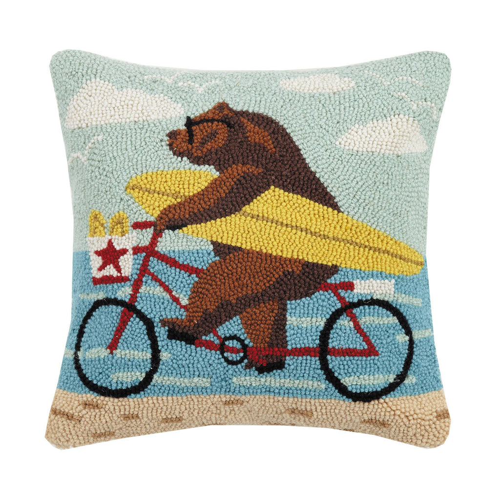 Hook pillow showing a bear in sunglasses riding a red bicycle with a yellow surfboard at the beach, sky and ocean in the background.