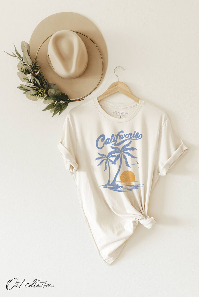 Woman wearing a cream t-shirt with a retro-style blue and gold “California” sunset graphic and palm trees.