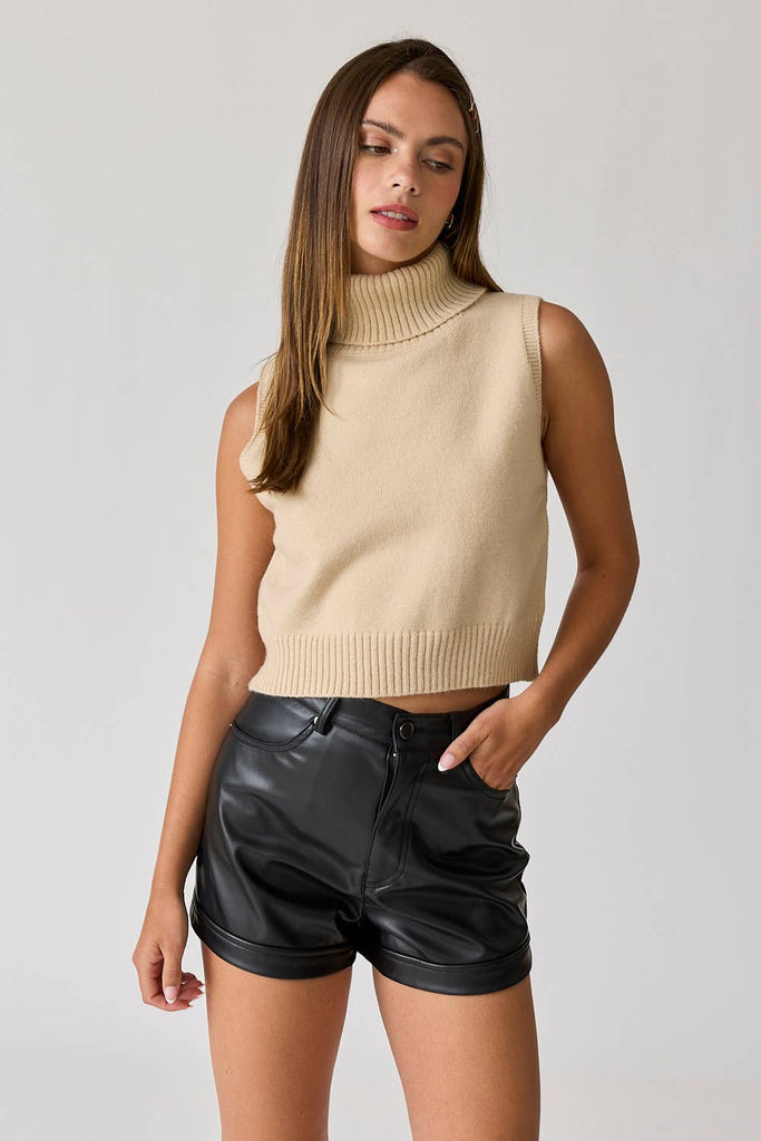 Woman wearing a beige sleeveless turtle neck knit cropped top with black leather shorts.