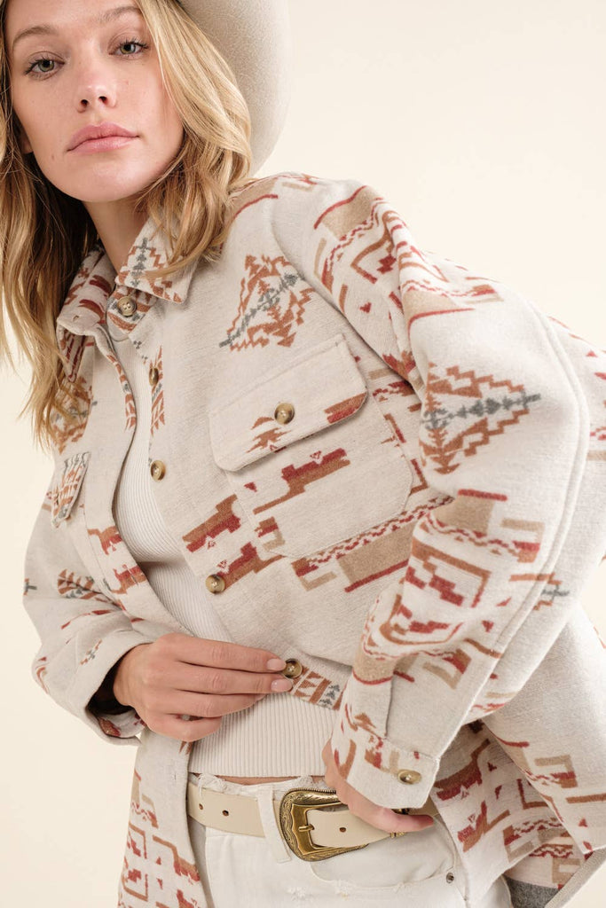 A woman wearing a beige Aztec-patterned jacket paired with a white tank top, distressed shorts, and a beige hat.