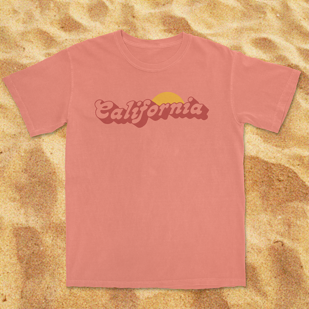 Vintage-inspired pink California T-shirt with a sun graphic on a sandy background.