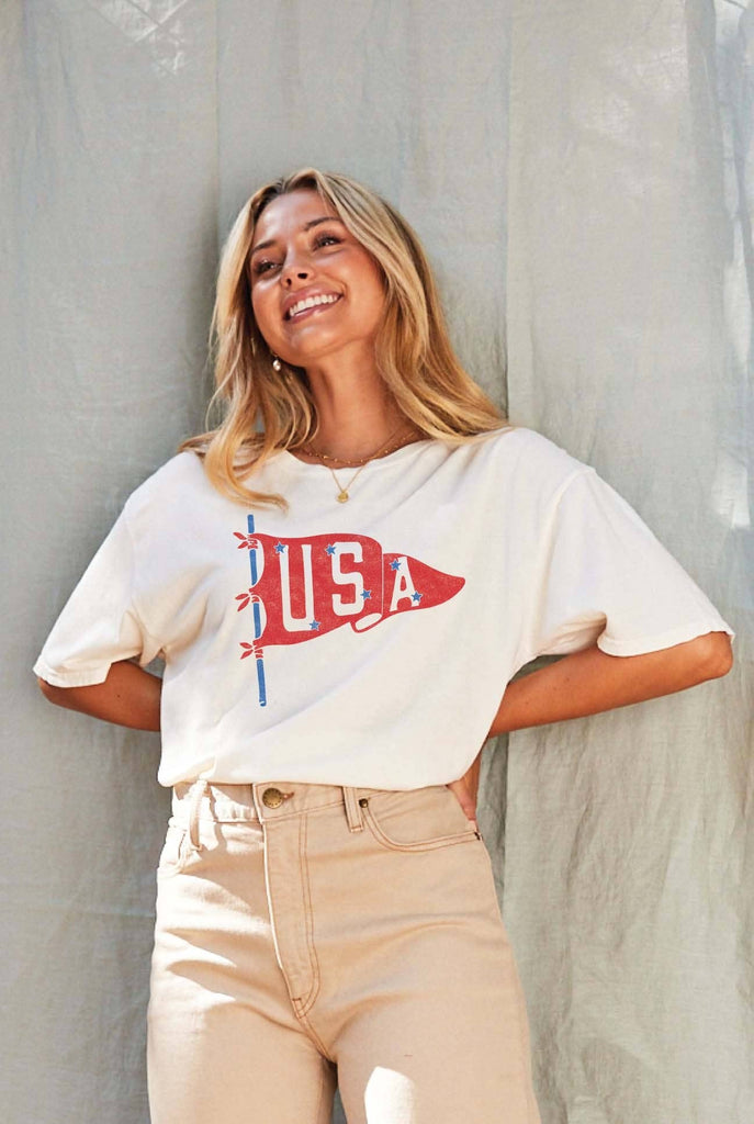 Woman wearing a cream-colored T-shirt with a vintage red USA pennant graphic, styled with beige high-waisted pants.