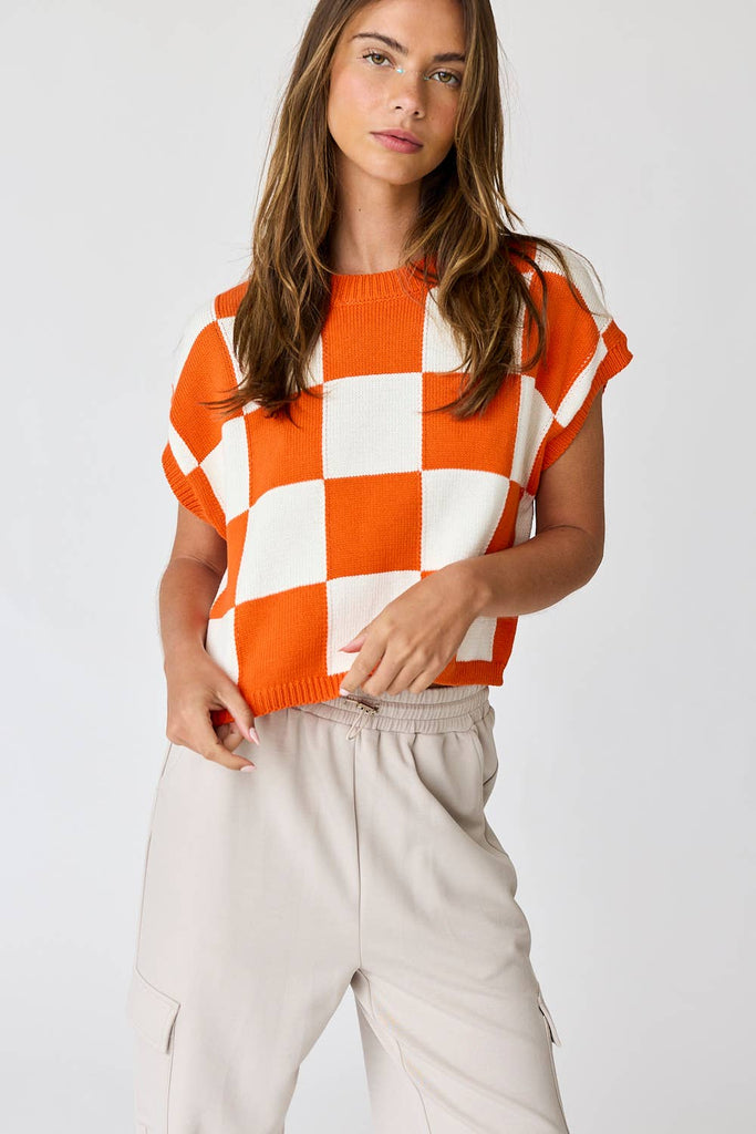 Woman wearing an orange and white loose-fit checker knit top with beige pants.