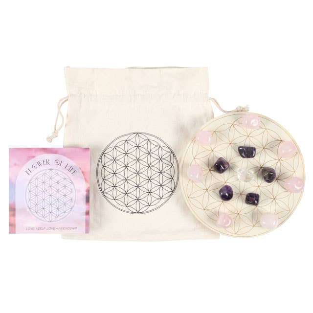 A Flower of Life crystal grid set featuring a wooden grid with a geometric flower of life pattern, arranged with various crystals including rose quartz, amethyst, and clear quartz. Surrounded by candles, flowers, and additional crystals on a black background.