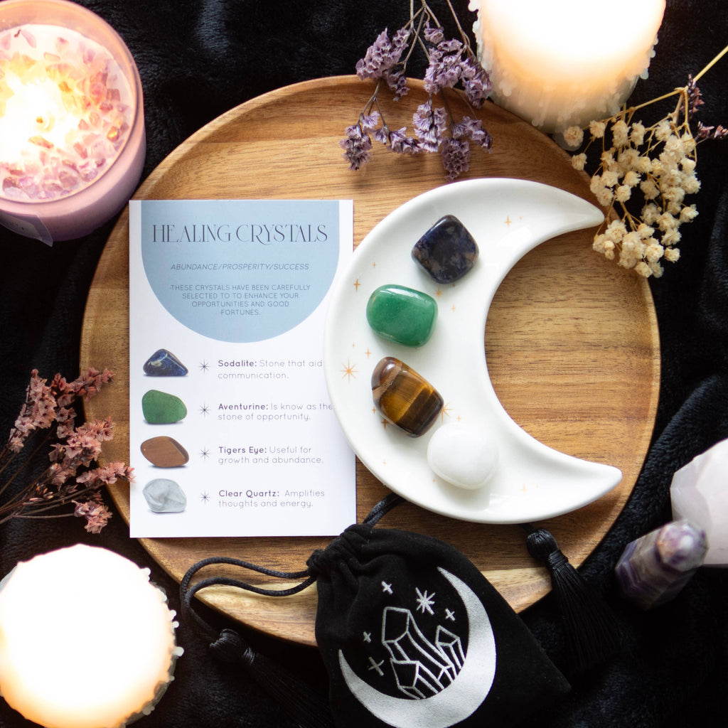 A success crystal set featuring a moon-shaped dish holding four stones (sodalite, aventurine, tiger’s eye, clear quartz), a black pouch with a crystal design, and a healing crystals information card.