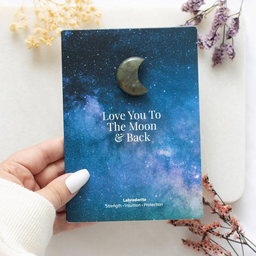 Hand holding a greeting card with a small labradorite crystal crescent moon and ‘Love You To The Moon & Back’ text.