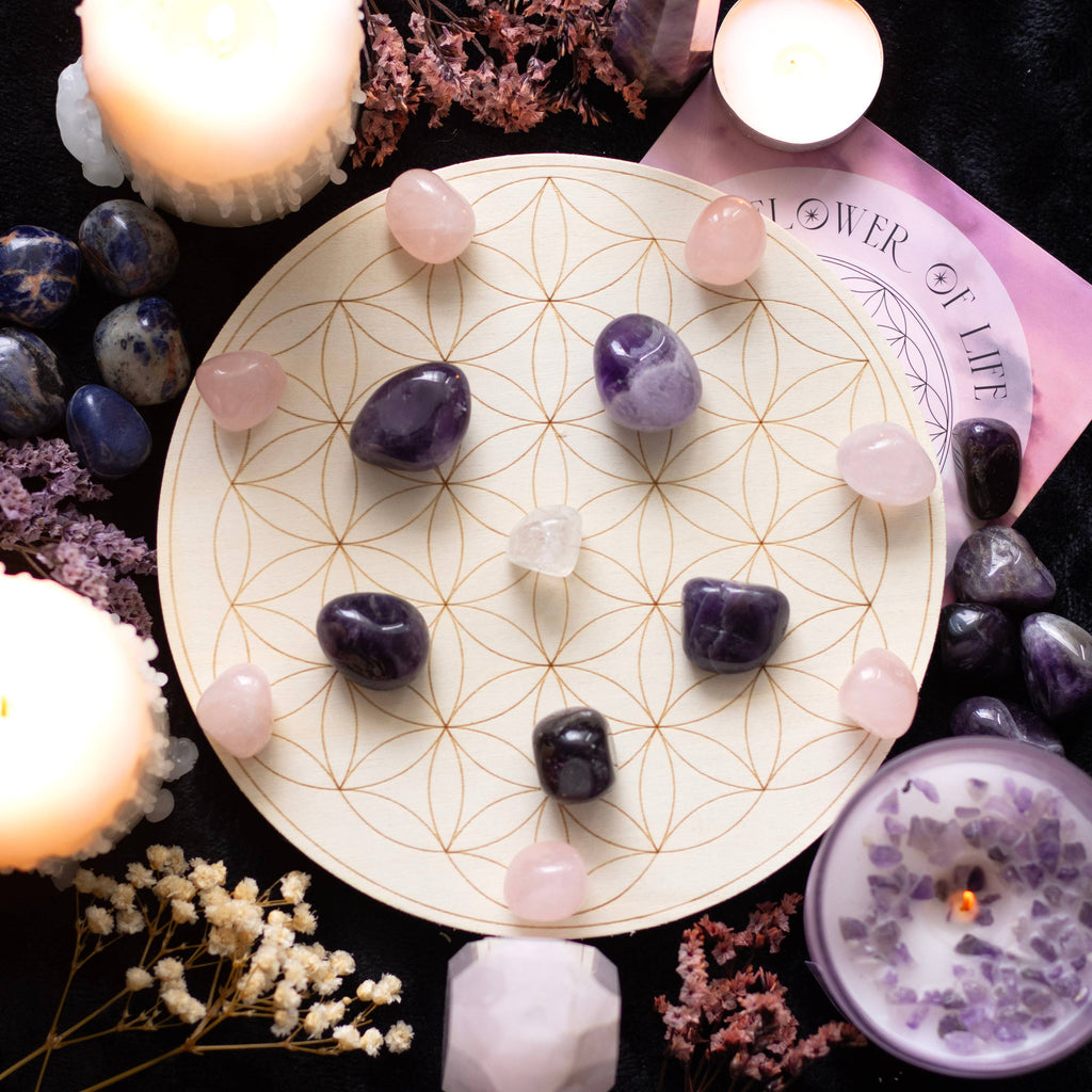 A Flower of Life crystal grid set featuring a wooden grid with a geometric flower of life pattern, arranged with various crystals including rose quartz, amethyst, and clear quartz. Surrounded by candles, flowers, and additional crystals on a black background.