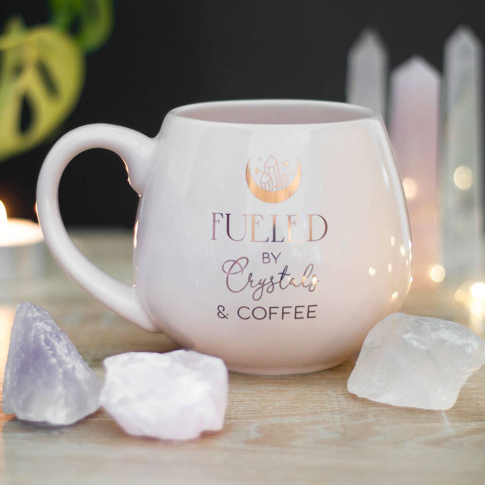 A stylish coffee mug with the text “Fueled by Crystals & Coffee,” surrounded by various crystals on a wooden table.