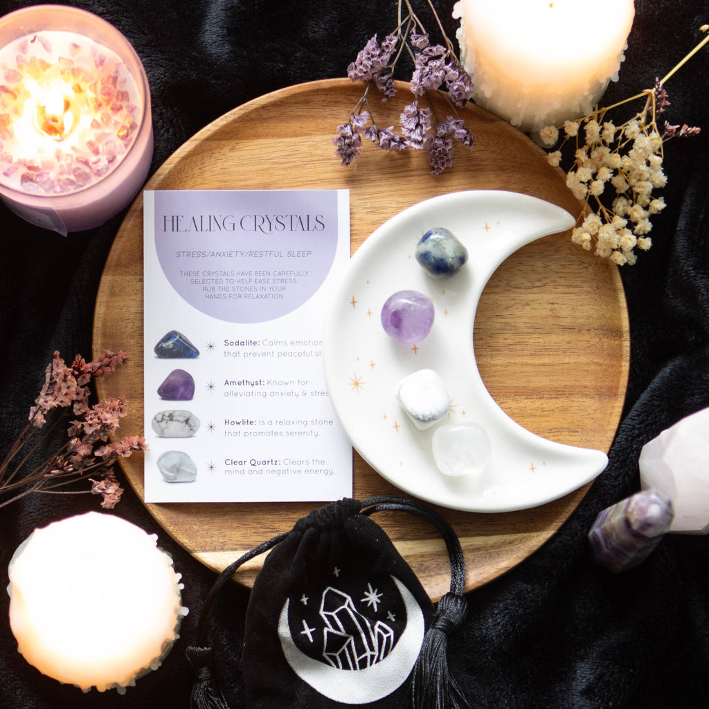 Stress Healing Crystal Set with Sodalite, Amethyst, Howlite, and Clear Quartz tumblestones on a crescent moon trinket dish, alongside a matching information card and printed drawstring pouch.